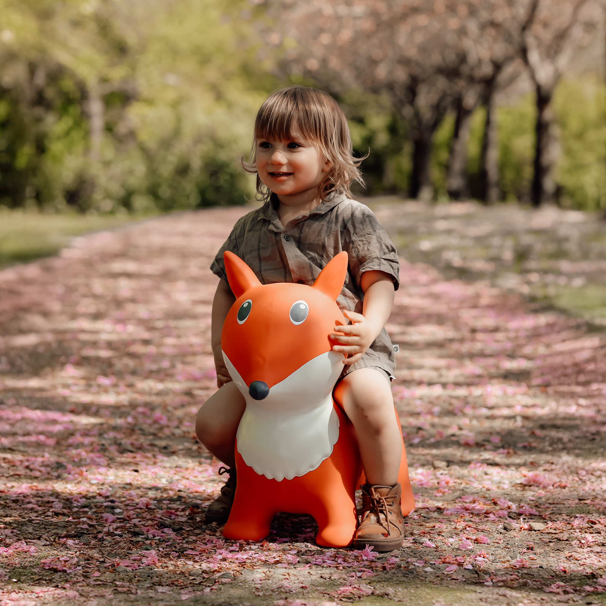 Niña montada en Lela, un zorro de juguete naranja y blanco que rebota, en un sendero cubierto de flores rosas – Encuentra a ese hipopótamo